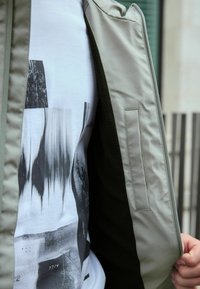 Personne portant une veste beige ouverte laissant apparaître un T-shirt blanc avec un motif géométrique abstrait noir et gris.