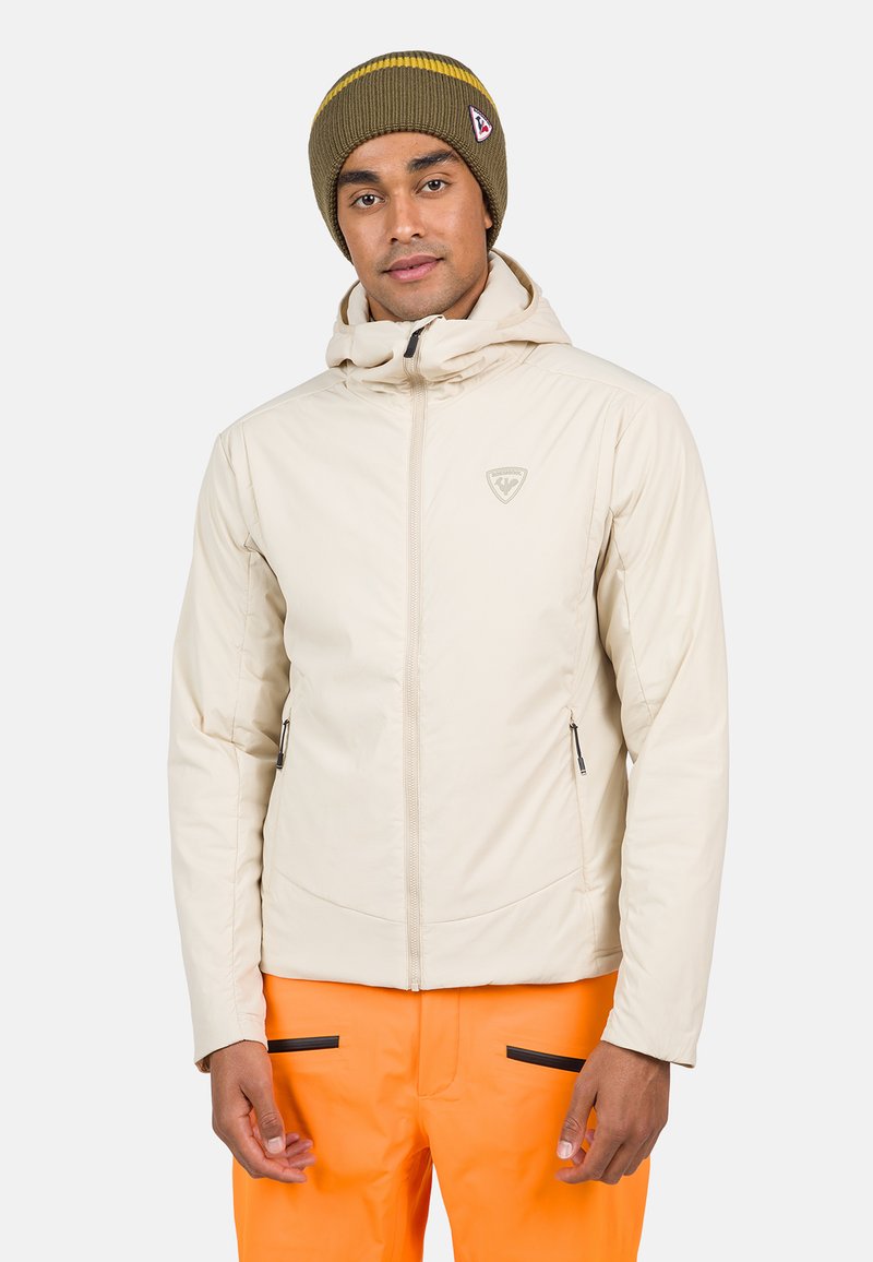 Veste isolante beige clair avec capuche, deux poches zippées et un logo sur la poitrine. Portée avec un pantalon orange vif et un bonnet noir.