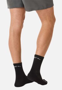 Schwarze Baumwollsocken mit einem gerippten Bündchen und einem kontrastierenden weißen Logo. Knöchellang, mit glatter Textur, getragen zu grauen Shorts.