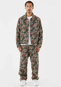 Jakke og bukser i mørkegrøn med blomsterprint, prydet med røde, lyserøde og cremede roser. Stoffet har en tekstureret overflade og er suppleret med hvide sneakers.