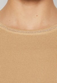 Top en tricot beige à côtes avec un col rond, présentant une texture lisse et un subtil fil brillant le long du col.