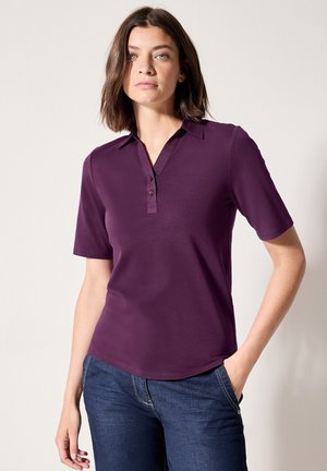 Camiseta polo corta de manga en color púrpura con cuello y tapeta de tres botones, fabricada en tejido suave, combinada con jeans oscuros de mezclilla.