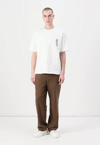 Fehér pamut póló rövid ujjakkal, bal mellkason fekete virágmintával, barna nadrággal és fehér sneakerrel párosítva.