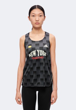 Mladá žena nosí čierne kárované tielko Adidas s nápisom „New York Together“ a čierne legíny na jednofarebnom pozadí.