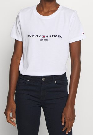 Personne portant un T-shirt blanc Tommy Hilfiger avec logo et texte "EST. 1985", associé à un jean bleu foncé.