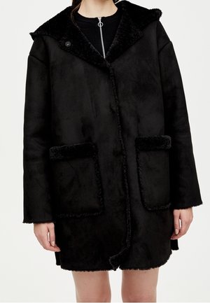 Schwarzer oversized Shearling-Mantel mit großen aufgesetzten Taschen, weitem Kragen und Knopfverschluss, getragen über einem schwarzen Top mit Reißverschluss.