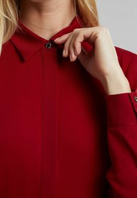 Czerwona bluzka z kołnierzykiem i zapięciem na guziki przy szyi. Gładki materiał o subtelnym połysku. Długie rękawy z mankietami zapinanymi na guziki.