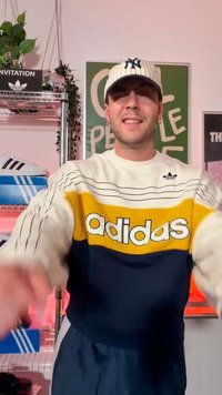 Νέος άνδρας που φοράει ριγέ πουλόβερ Adidas και καπέλο New York Yankees, στέκεται σε ένα δωμάτιο με αθλητικά παπούτσια και φυτά σε ράφια πίσω του.