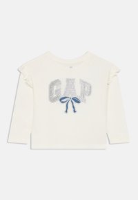Chemise blanche à manches longues avec un logo "GAP" pailleté et un motif de nœud bleu. Détails à volants délicats sur les épaules. Matière en coton doux.