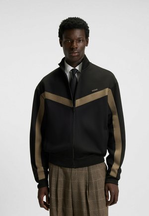 Sort lynlåsjakke med beige chevron- og ærmestriber, båret over hvid skjorte og sort slips, parret med brune rutede bukser.
