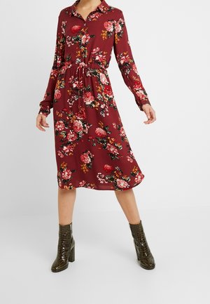 Woman wearing a maroon floral button-down midi dress with long sleeves and tied waist, paired with dark green textured ankle boots.