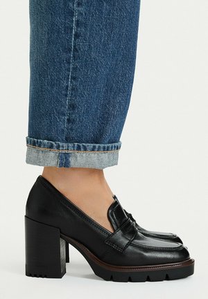 Fod iført sort læder loafer med blokhæl parret med blå opkrogede denimjeans på hvid baggrund.