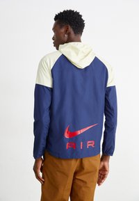 Jūras spēku un gaiši krēmkrāsas divu toņu vēja jakas ar kapuci. Iespējas ietver lielu sarkanu Nike AIR logotipu aizmugurē, elastīgas piedurknes un vieglu materiālu.