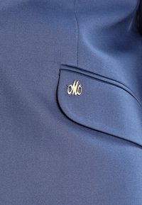 Námořnická modrá látka zachycující detail kapsy se zlatým akcentem s propletenými písmeny "M" a "O". Hladká textura, přiléhavý střih.