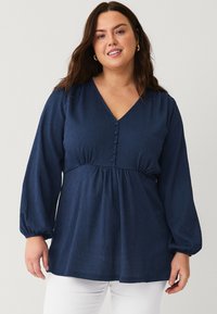 Navy blue long-sleeve blouse with a V-neck, button details, gathered waist, and textured fabric. Paired with white pants.