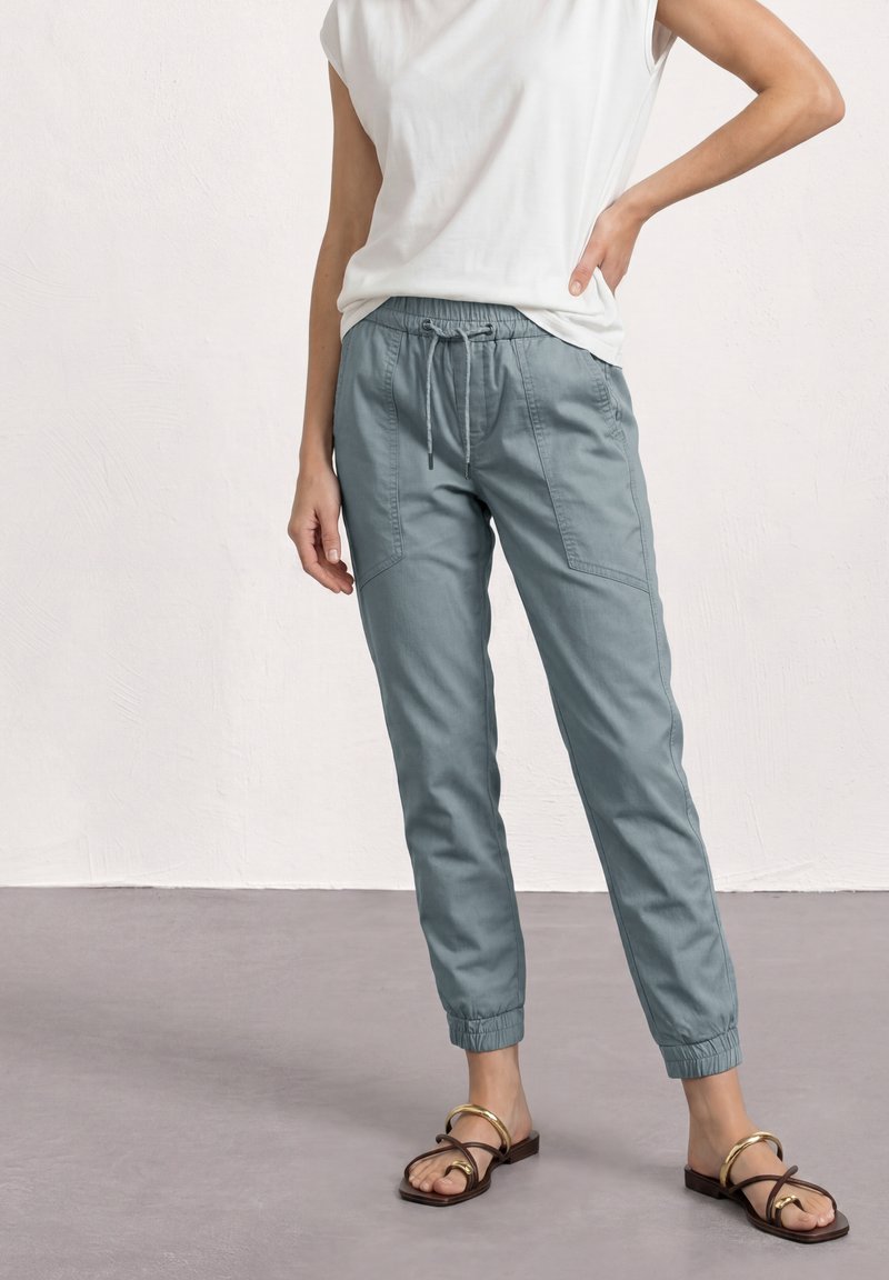Personne portant un pantalon jogger élastique bleu clair aux chevilles, un débardeur blanc et des sandales marron avec des accents dorés, debout sur un sol gris.