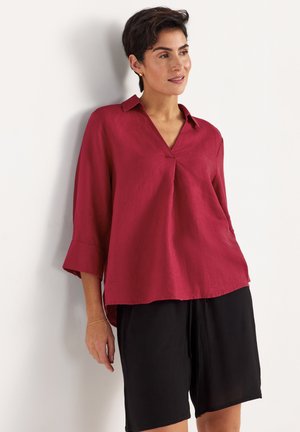 Kobieta opierająca się o ścianę, ubrana w luźną czerwoną bluzkę z kołnierzykiem i rękawami do połowy przedramienia oraz czarne krótkie spodenki sięgające do kolan.