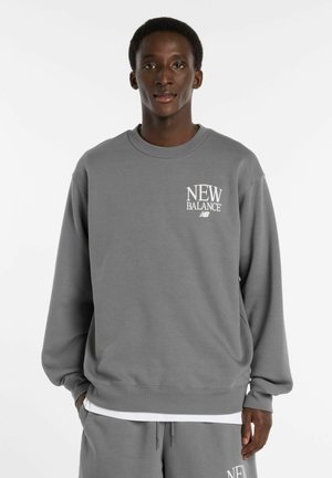 Mann trägt einen grauen New Balance Pullover und passende Hose, steht mit den Händen in den Taschen vor einem weißen Hintergrund.