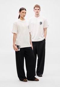 Dve osebi nosita svetlo bež prevelike majice s potiskom spredaj, združene z ohlapnimi črnimi hlačami. Ena drži prozorno torbo.