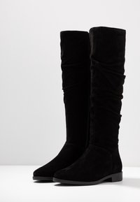 Czarne zamszowe kozaki sięgające kolan o luźnym kroju, z ostro zakończonym noskiem i niskim, grubym obcasem, z gładką teksturą i subtelnymi detalami w postaci przeszyć.