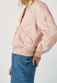 Femme portant une veste bomber rose clair avec fermeture éclair et poches à rabat, associée à un jean en denim bleu taille haute sur un fond uni.