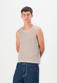 Top de tirantes de punto acanalado beige, con cuello redondo y amplias sisas, combinado con jeans de mezclilla azul a rayas con bordado rojo en el bolsillo.