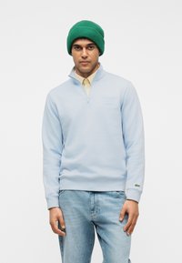Homme portant un bonnet en laine vert, un sweat-shirt à fermeture éclair partielle bleu pâle sur une chemise beige, et un jean bleu clair, debout devant un fond blanc.