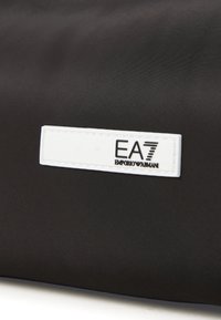 Borsa nera in nylon con superficie testurizzata e un'etichetta rettangolare in pelle bianca con logo marchiato in nero, "EA7 Emporio Armani."
