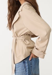 Veste beige ceinturée avec des manches longues, des revers à clou et des poches latérales. Le tissu semble lisse et léger, associé à un jean en denim bleu.