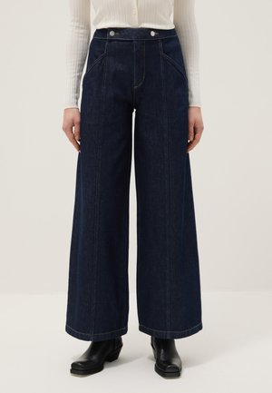 Wijd uitlopende donkere spijkerbroek met knoopdetails aan de voorkant, gedragen met zwarte enkellaarzen en een witte geribbelde top met lange mouwen.