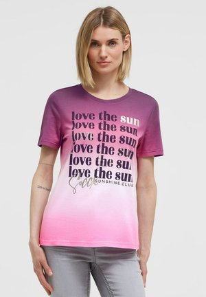 Woman with short blonde hair wearing a purple to pink ombre t-shirt with repeated "love the sun" text and light gray pants.