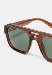Ochelari de soare din plastic maro cu lentile verzi, design cu un cadru unghiular, decupaje proeminente pe laterale și detalii subtile cu logo în față.