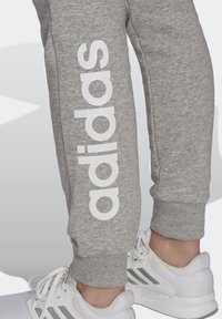 Grijze trainingsbroek met een witte "adidas" logo langs de zijkant. Geribbelde boorden en een zachte textuur, gecombineerd met witte sportschoenen met grijze accenten.