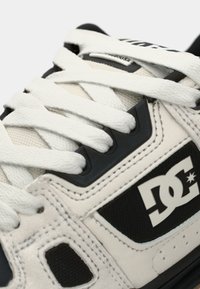 White sneaker with black accents, featuring a textured upper, prominent logo, and flat white laces. Close-up view of materials and design details.