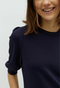 Maglietta navy a maniche corte con scollo rotondo e dettagli di maniche svasate, realizzata in tessuto morbido e liscio. In primo piano sulla parte superiore del corpo.