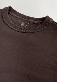 Pruun triiksärk, millel on sooniline ümar kaelus ja tekstureeritud kangas. Sildil on selgelt nähtav kaubamärgi nimi "NEXT".