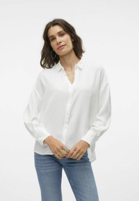 White button-up shirt with a v-neckline, long sleeves, relaxed fit, made from textured fabric. Paired with blue denim jeans.