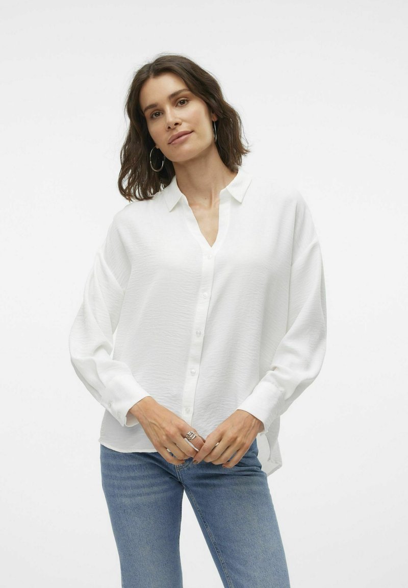 White button-up shirt with a v-neckline, long sleeves, relaxed fit, made from textured fabric. Paired with blue denim jeans.