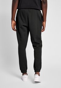 Sorte sweatpants lavet af blødt stof, med elastiske cuffs og en afslappet pasform. Der er en lomme på venstre side. Bæres med hvide sneakers.