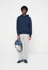 Felpa navy, pantaloni tuta grigi, borsa blu con logo Adidas, sneaker navy con tre strisce, vestibilità rilassata, materiale in cotone, design casual.