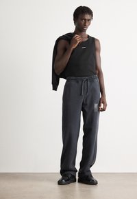 Black sleeveless tank top with "HUGO" logo, paired with dark gray sweatpants featuring an elastic waistband and side pockets. Black sneakers.