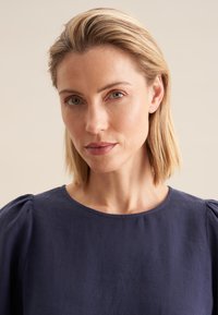 Top in lino blu navy con maniche corte a sbuffo, scollatura rotonda e una sottile texture. Ritratto ravvicinato che mette a fuoco il design del capo.