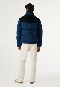 Temno modra pufasta jakna s prešano teksturo, kontrastnimi temnimi rameni in visokim ovratnikom; kombinirana s kremnimi hlačami in sproščenimi čevlji.