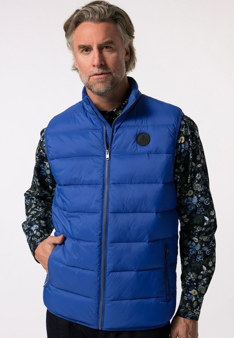 Blå vadderad väst med dragkedja, sidofickor och en rund svart logotyp-patch. Bärs över en mörk blommig mönstrad skjorta.