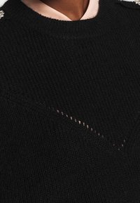 Pull en tricot noir avec une finition texturée, présentant un motif en V subtil et orné de décorations sur les épaules.