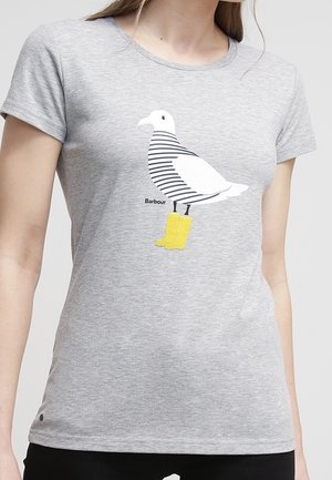 Vrouw die een grijs t-shirt draagt met een witte meeuw grafisch gestreept als een zeeman, staand in gele laarzen, met de tekst "Barbour".