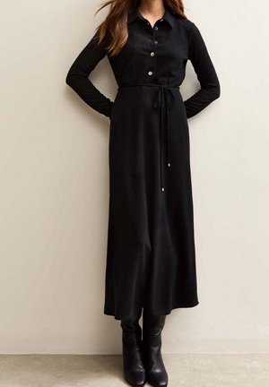 Femme portant une longue robe noire côtelée boutonnée avec une ceinture à la taille et des bottines noires, debout devant un mur clair uni.