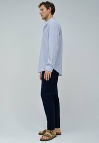 Camisa de manga larga a rayas azul claro y blanca con cuello abotonado, combinada con pantalones azul marino y sandalias marrones. Fondo liso.