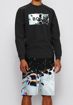 Man wearing black long-sleeve shirt with holographic "BOSS" logo and matching black, white, and multicolor abstract patterned shorts.