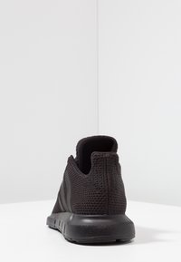 Svart atletisk sko med en texturerad mesh-övre, gummisula, vadderad krage och minimalistisk design. Randiga accenter på sidan.